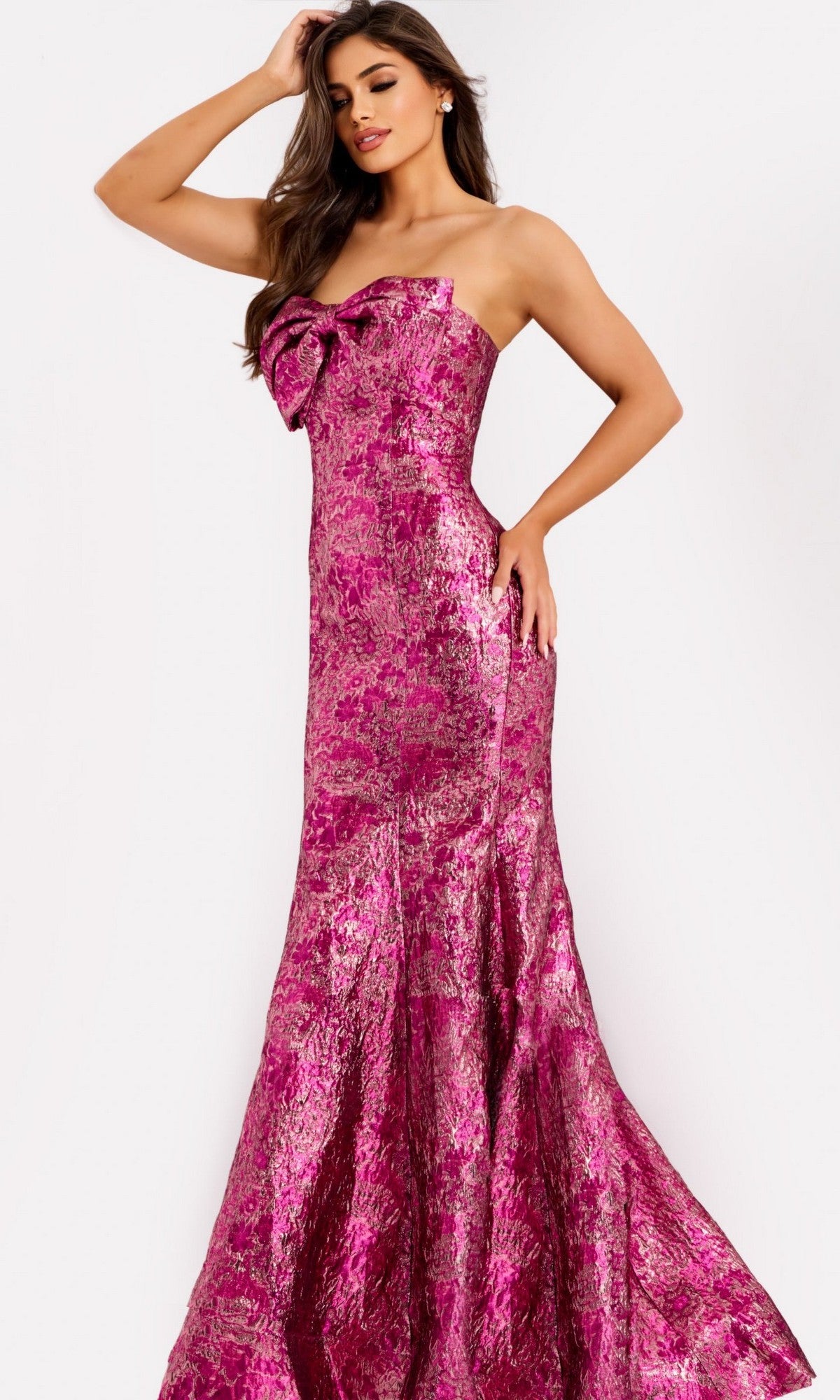 A woman poses against a plain white background wearing the Jovani 49163 strapless long metallic prom dress, featuring a floor-length mermaid skirt and a large bow on the bodice.